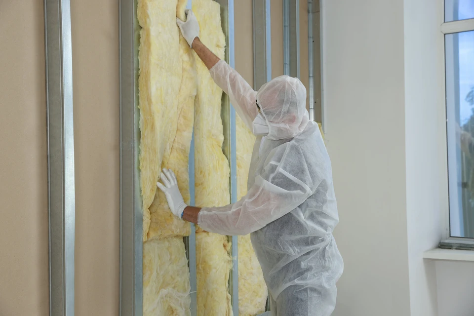 Intérieur de bâtiment en construction, ouvrier en combinaison de protection blanche et gants installant de la laine de verre jaune entre montants métalliques d'une cloison