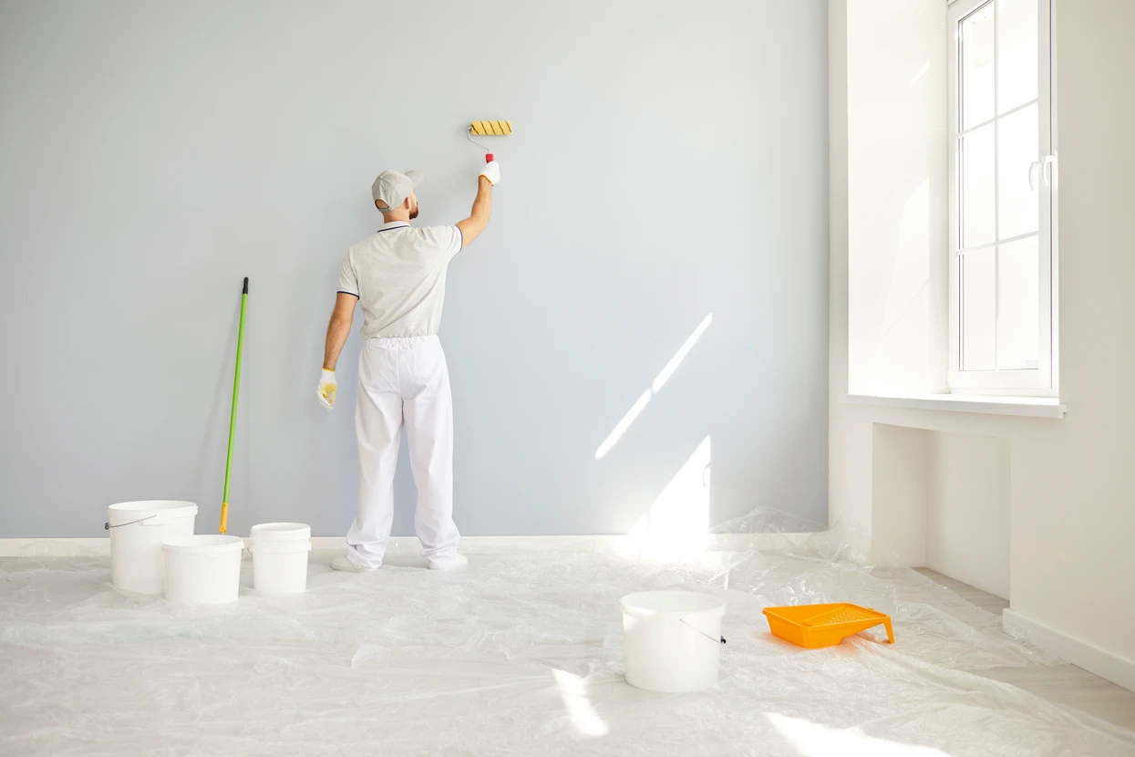 Intérieur d'une pièce en cours de rénovation avec mur partiellement peint en jaune vif, échelle escabeau en bois positionnée au centre, pots de peinture et pinceaux disposés sur parquet stratifié