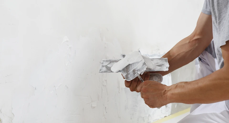 Homme mûr en salopette jaune appliquant de la peinture blanche sur un mur intérieur à l'aide d'un rouleau à manche télescopique rouge